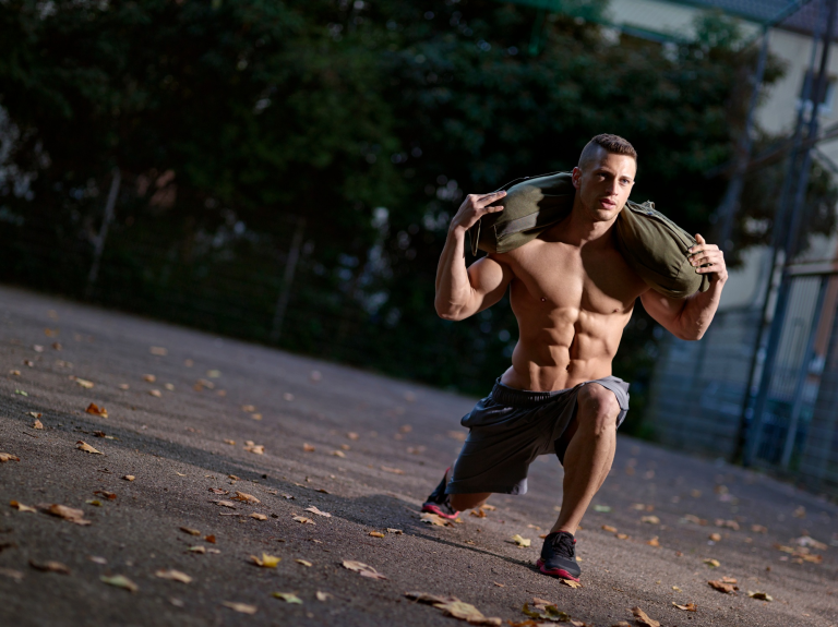 Muscly Young Man Training With Sandbag On Sports Field